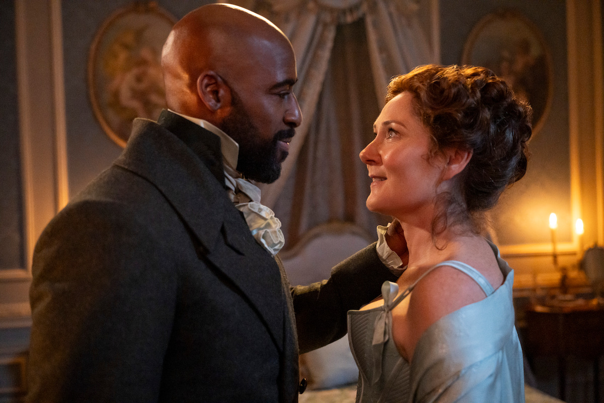 A man and woman in period clothing gaze at each other warmly in an elegant, candlelit room with ornate decor and soft lighting, suggesting a romantic or dramatic moment in a historical setting.