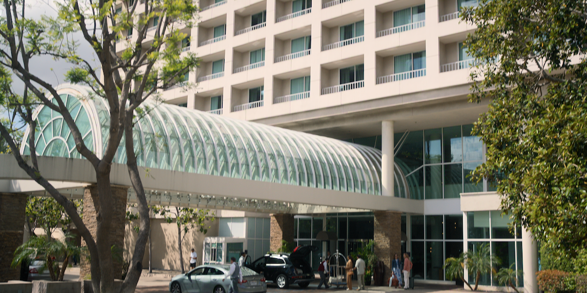 Modern hotel exterior with a glass covered walkway, people and cars at the entrance, surrounded by trees and greenery on a sunny day.