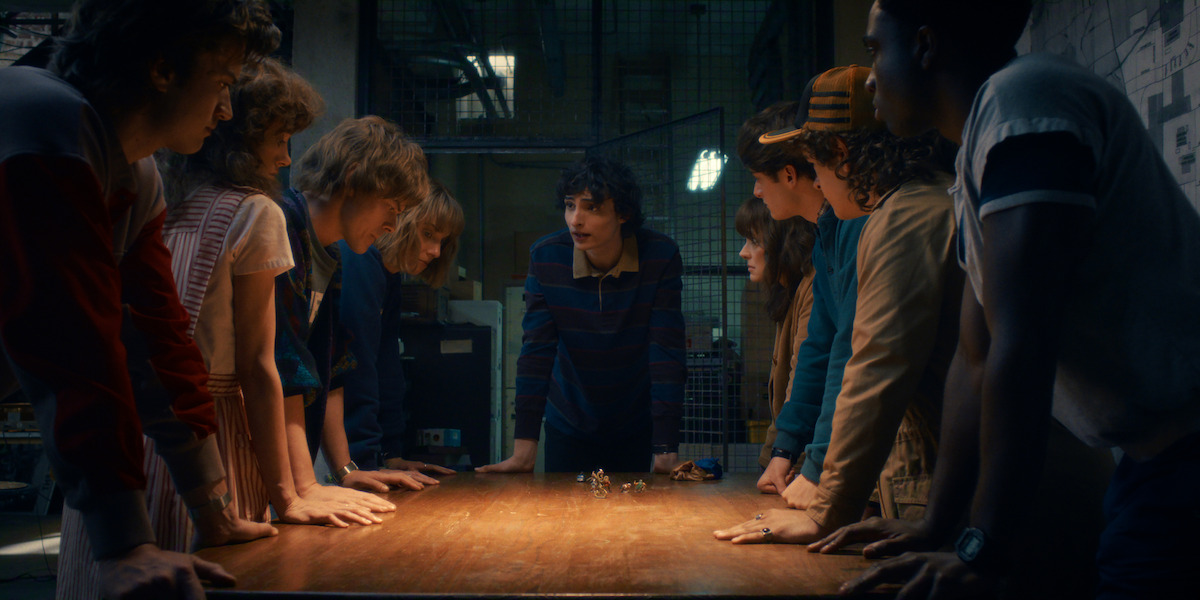 Five teenagers gather in a dim, basement-like room with a cluttered desk, chairs, a large map on the wall, and soft, dramatic lighting, suggesting a tense group discussion or planning session.