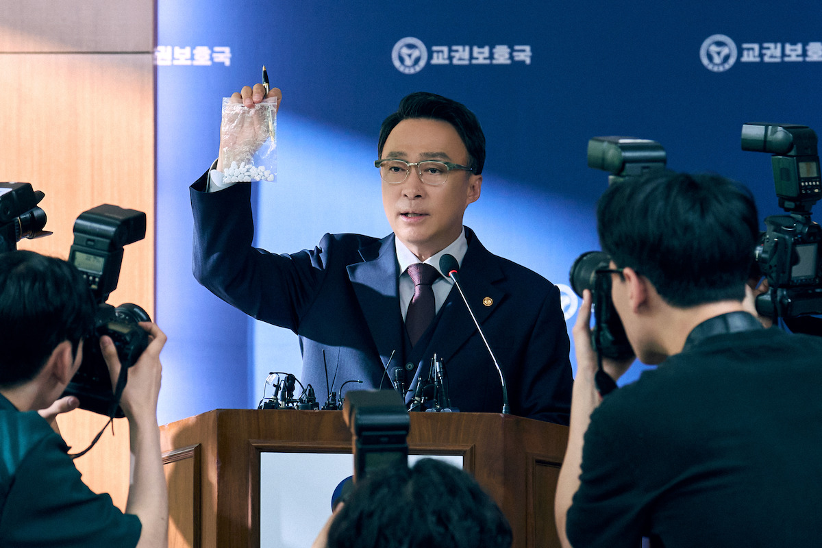 A gentleman in glasses and a suit holds up a clear zipped bag filled with pills while addressing a group reporters at a press conference.A gentleman in glasses and a suit holds up a clear zipped bag filled with pills while addressing a group reporters at a press conference.