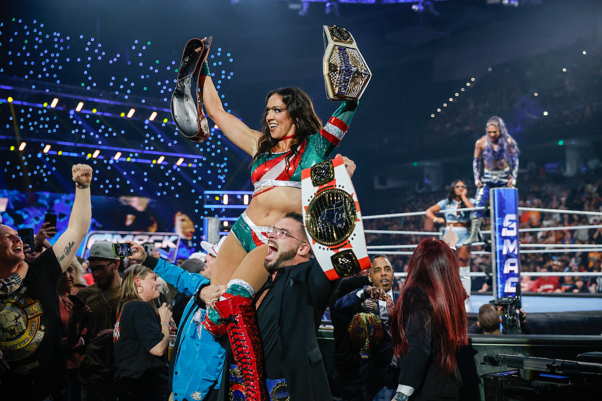 Female wrestler celebrates in a packed arena, holding championship belts while lifted by fans. The environment is energetic, with lights and a wrestling ring, capturing a triumphant, festive mood.