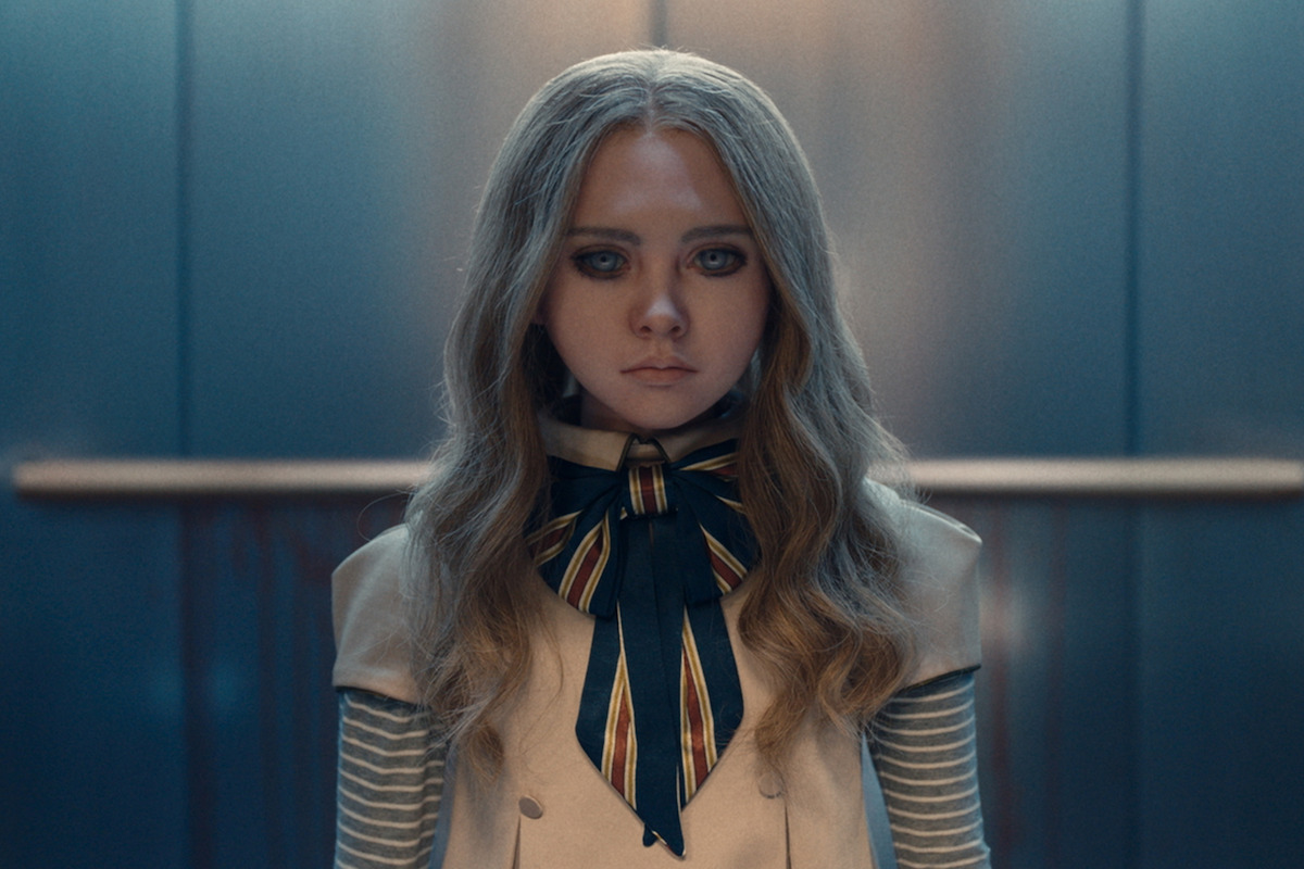 A robotic girl with long blond hair, blue eyes, and a striped bow stands in a modern, dimly lit elevator with metal walls, giving a neutral, slightly eerie expression.