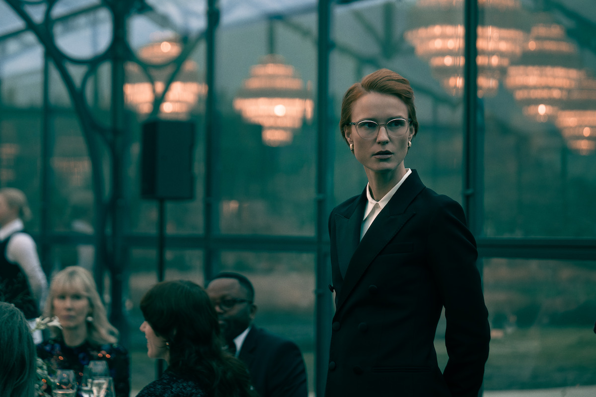 Amanda Collin wears an updo, glasses, and suit and while looking off into the distance with a serious expression.