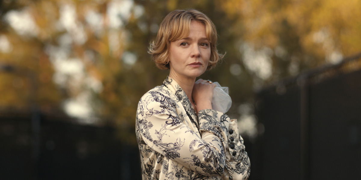 Woman with short blond hair wearing a patterned jacket stands outdoors in front of a blurred background of autumn trees, holding a face mask in her hand and looking thoughtfully into the distance.