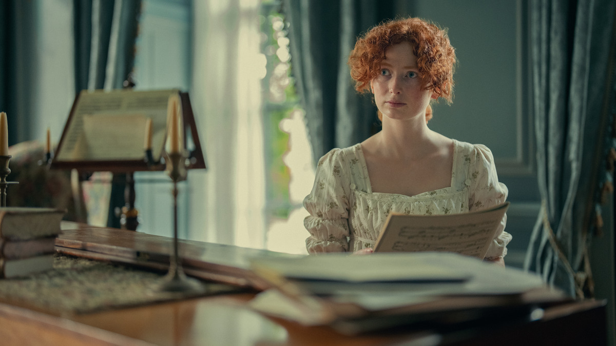 A young woman with curly red hair in a vintage dress sits at a piano in a softly lit, elegant room with candles and sheet music, holding a music sheet and looking contemplative.