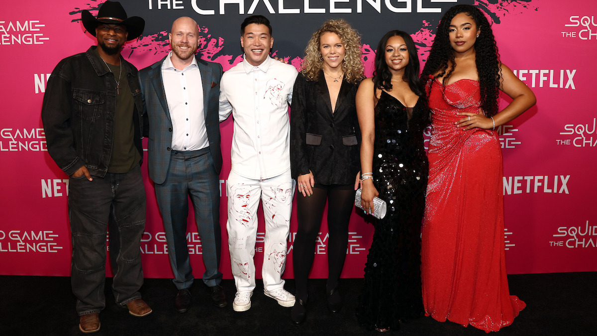 Six people dressed formally pose on a red carpet at the "Squid Game: The Challenge" event, standing in front of a branded backdrop. The atmosphere is lively and celebratory.