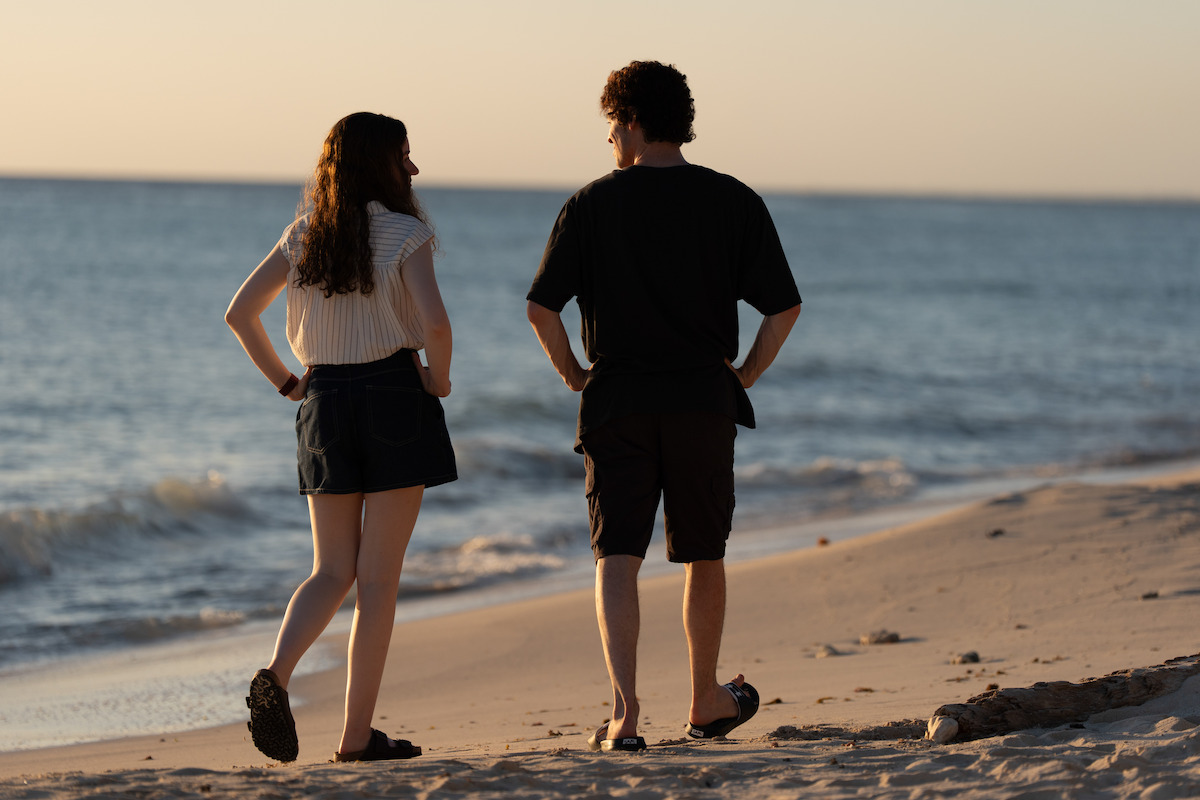 Two people walking along a sandy beach at sunset, facing the sea and each other, with gentle waves rolling in the background and soft evening sunlight illuminating the shoreline.