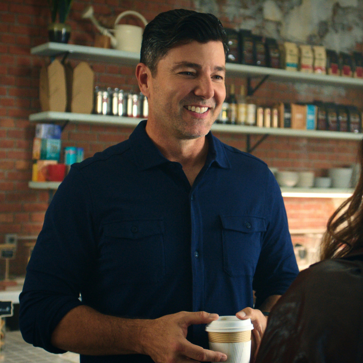 Man in a blue shirt smiling and holding a coffee cup in a cozy, modern coffee shop with exposed brick walls, shelves with coffee bags, mugs, and a plant in the background.
