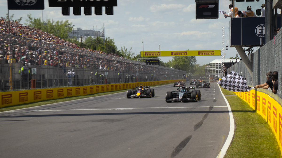 Formula 1 race cars cross the finish line on a sunny day at a racetrack, with a waving checkered flag, large crowd in grandstands, Pirelli banners, and trackside structures in the background.