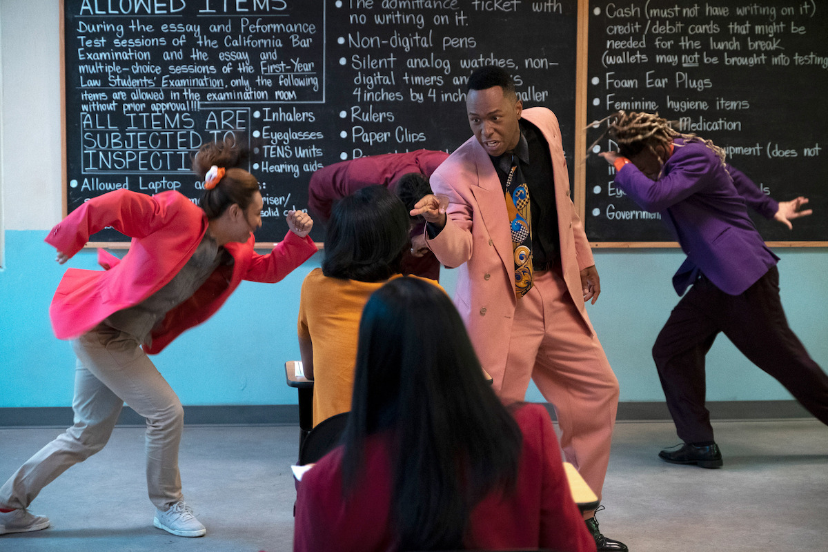 a teacher dress in a pastel suit sings in front of a blackboard filled with instructions