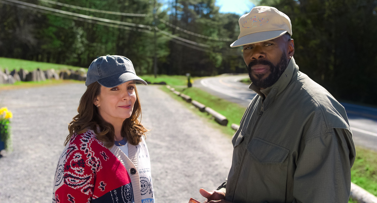 Two people wearing casual clothes and hats stand on a sunny roadside with green trees and plants in the background, suggesting a relaxed outdoor setting in nature.