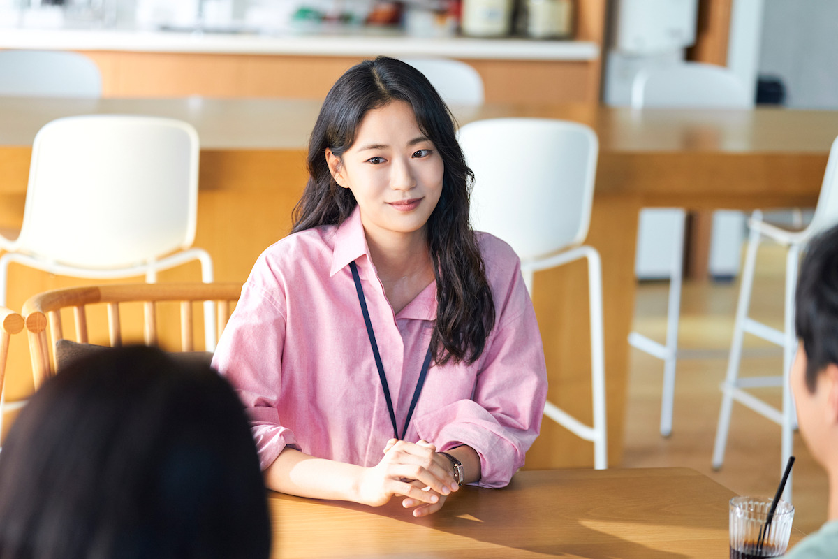 Woman in a pink shirt sits at a wooden table, smiling and facing two people in a bright, modern room with white chairs and a kitchen in the background.