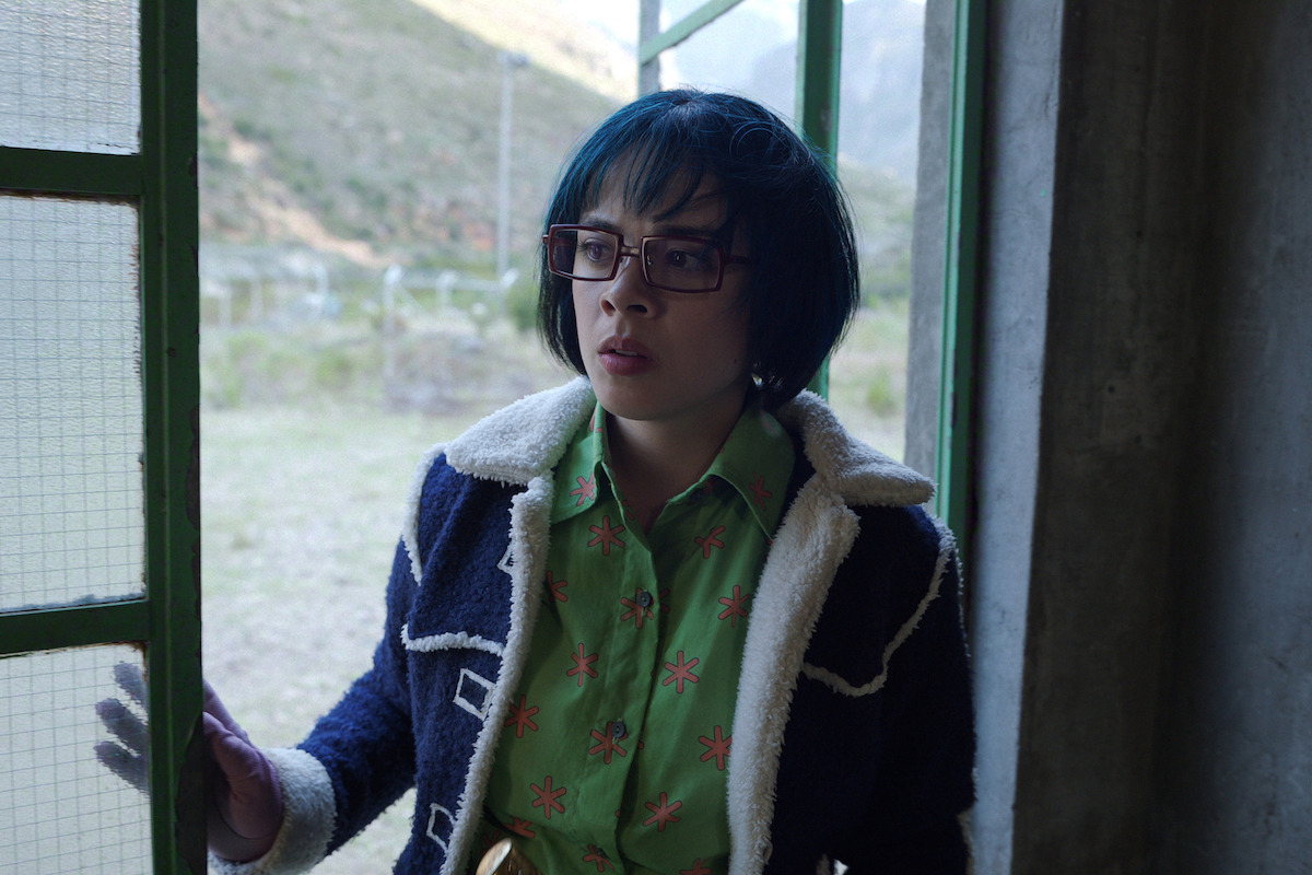 Person with short dark hair and glasses stands in a doorway, partially inside a building with concrete walls and glass door, looking outside at a rural landscape with hills in the background, wearing a patterned green shirt and jacket.
