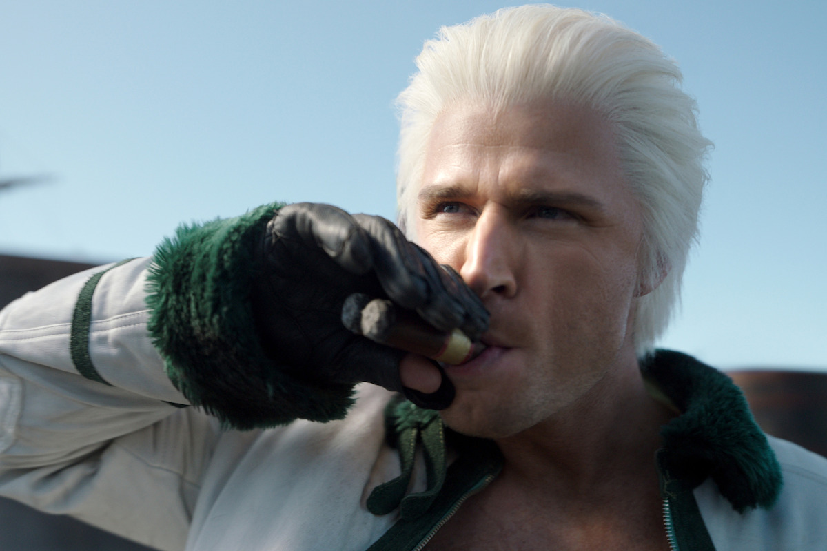 A man with white hair and a white jacket with green trim drinks from a metal flask outdoors in daylight, wearing a black glove and looking intense, with blue sky in the background.