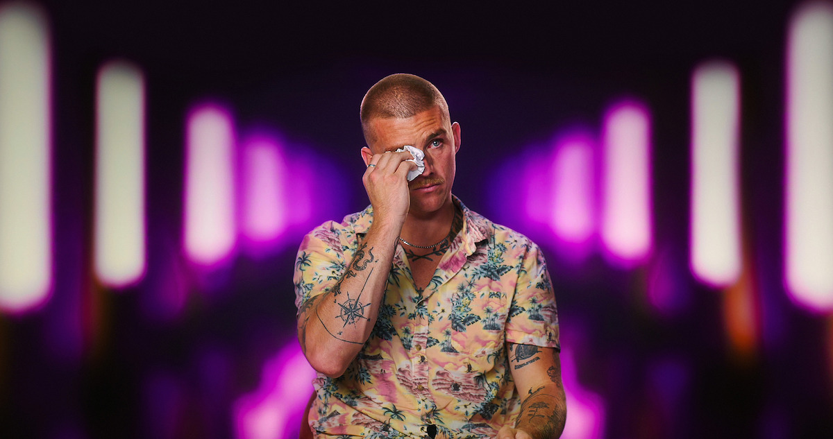 A man in a floral shirt wipes his eye, sitting in front of a blurred background with vertical neon purple and white lights, giving a dramatic nightclub or studio environment.