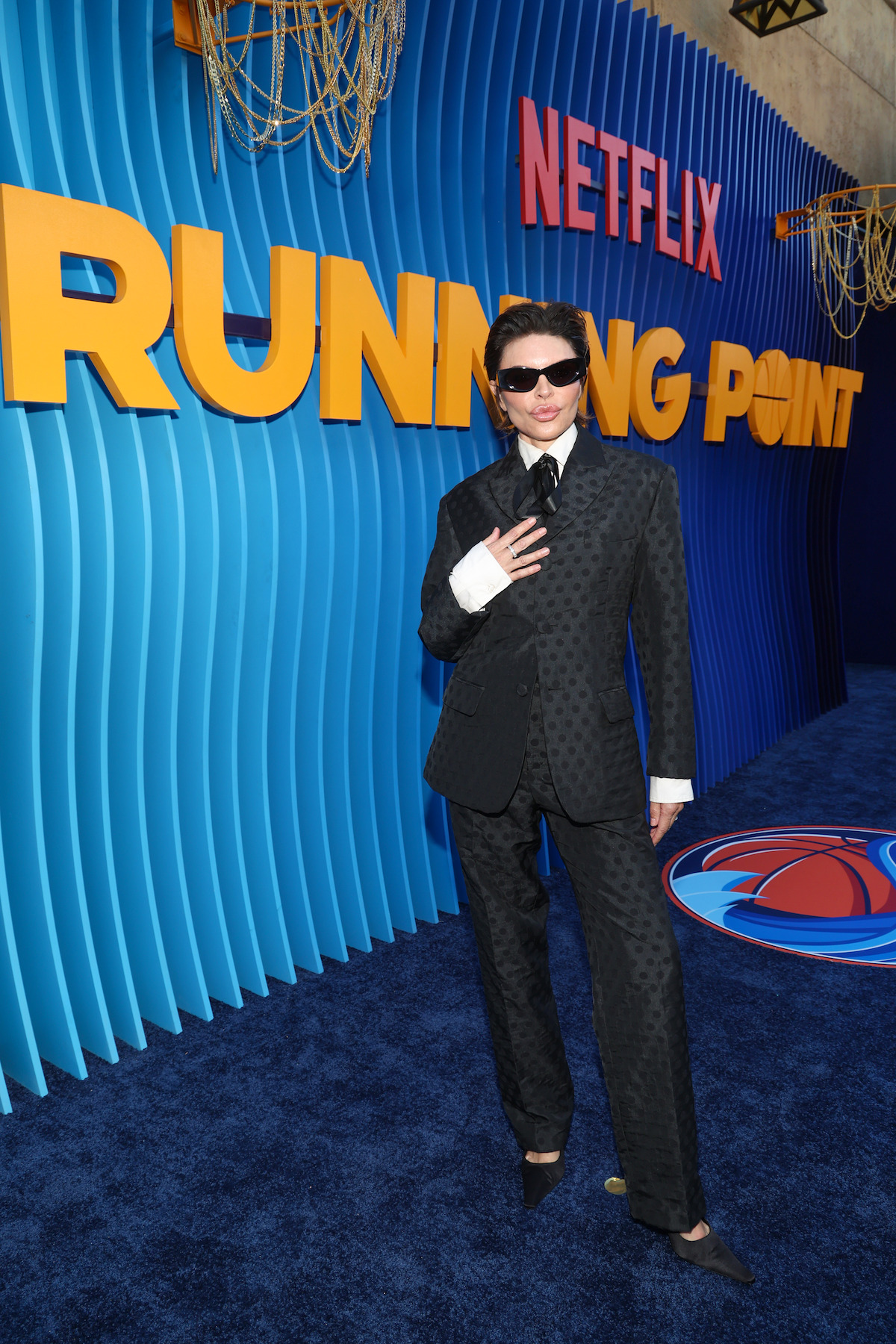 Person in a dark patterned suit and sunglasses poses on a blue carpet in front of a large Netflix "Running Point" promotional backdrop with bold yellow lettering and decorative lighting fixtures, creating a formal event atmosphere.