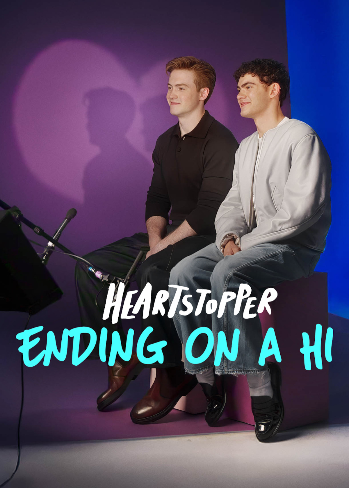 Two young men sit side by side on a purple bench in a studio with purple and blue lighting, microphones in front, smiling, with Heartstopper: Ending on a Hi text overlayed.