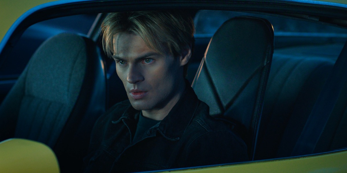Young man with blond hair sits in the driver's seat of a car at night, looking serious and focused; the car has a dark interior and yellow exterior, illuminated by blue-toned lighting.