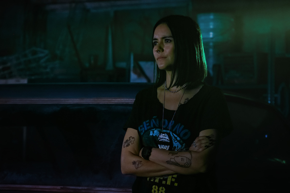 A woman with dark hair and tattoos stands with arms crossed in a dimly lit, industrial setting with greenish-blue lighting, next to a dark vehicle and surrounded by metal objects and shadows.