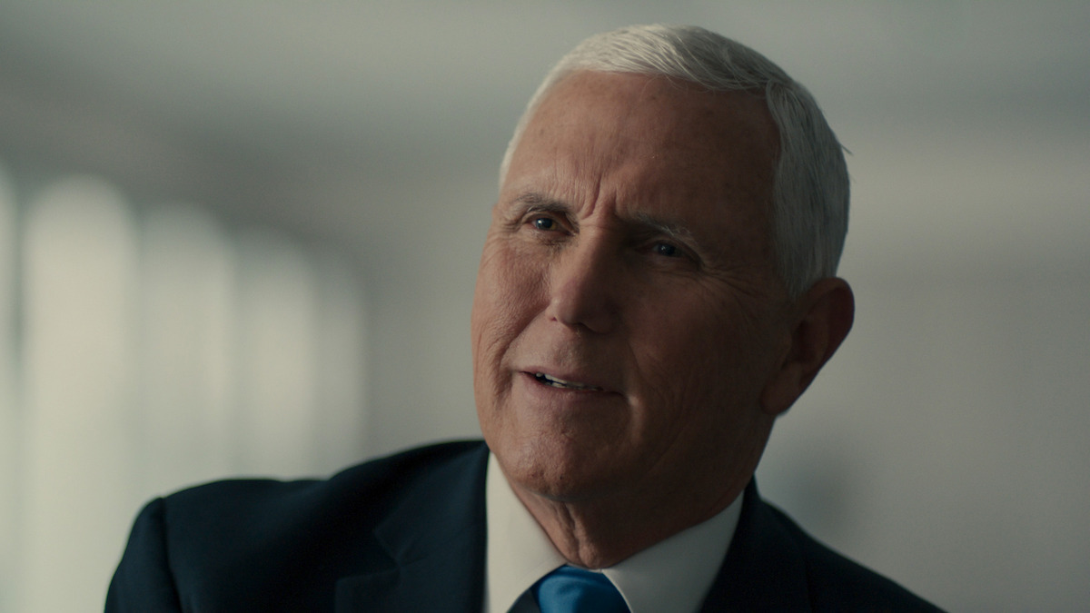 Older man with short gray hair wearing a suit and blue tie, seated indoors in a softly lit room with blurred windows in the background, looking slightly off camera with a thoughtful expression.