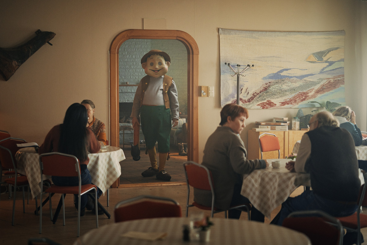 Person in a large Pinocchio costume stands in a doorway of a small café, where people sit at tables with checkered tablecloths, chatting and drinking in a cozy, warmly lit interior with wall art and wooden decor.