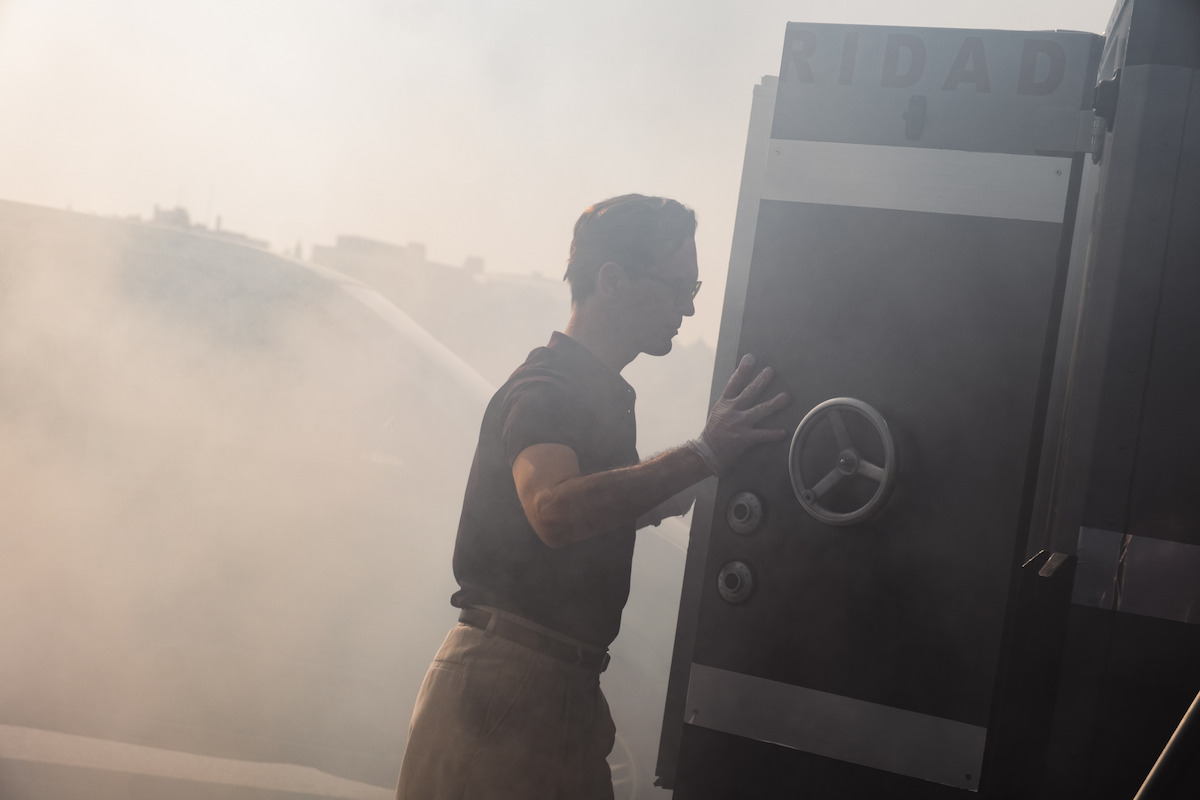 Man in sunglasses and dark shirt pushing a large metal door or vault in a smoky outdoor environment, with a vehicle and indistinct cityscape visible in the background.