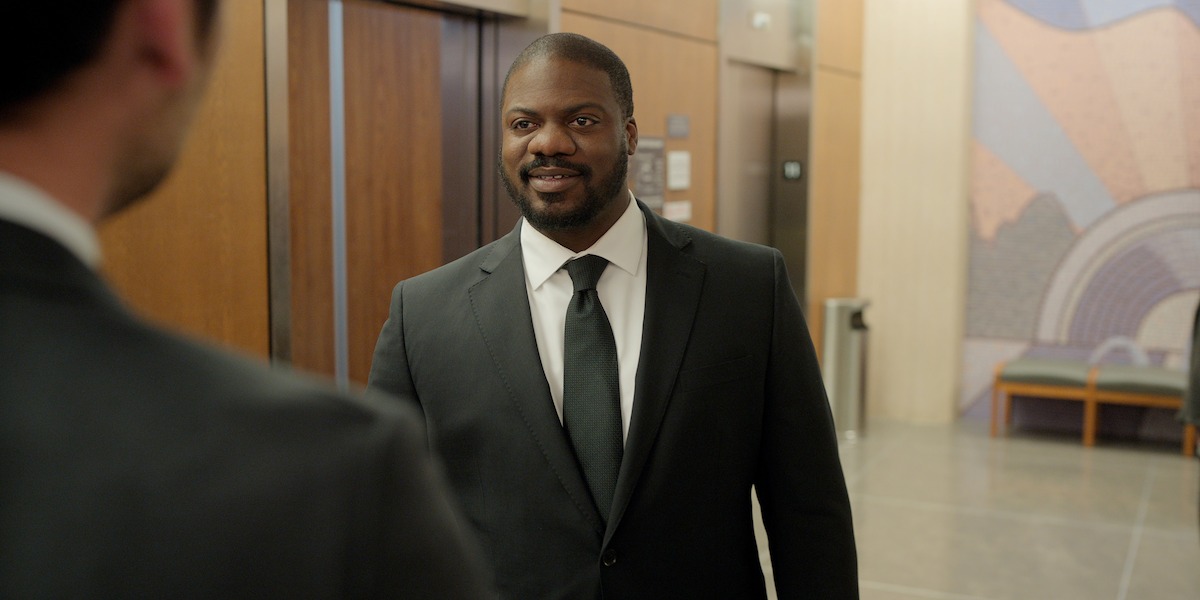 A man in a suit smiles while standing in a modern office lobby with elevators, art on the wall, and a bench in the background.