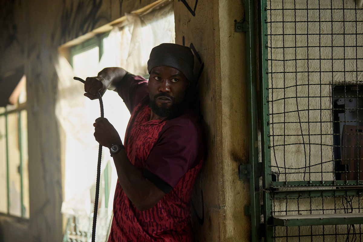 Man wearing a red shirt and head wrap cautiously holding a rope, standing against a wall in a dim, graffiti-covered, abandoned building with metal gates and sunlight streaming through windows.