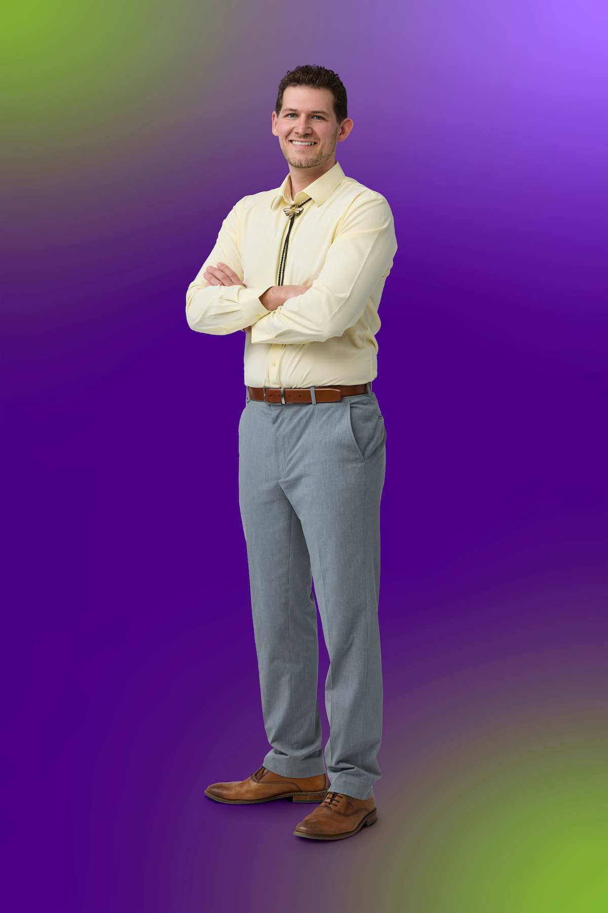 Smiling man in a yellow shirt and tie with gray pants stands with arms crossed on a gradient purple and green background, posing confidently in a studio setting.