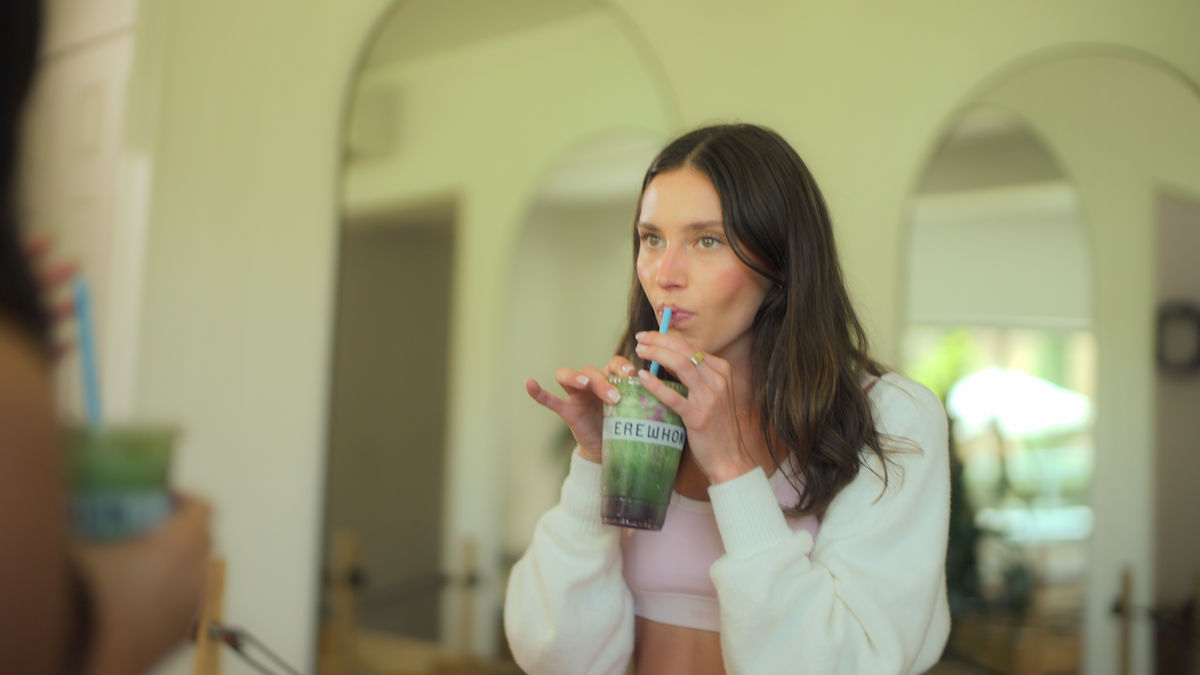 Woman in casual indoor setting sipping a colorful drink through a straw, standing in front of arched mirrors. Bright, soft lighting creates a relaxed and cozy atmosphere. Another hand holding a similar drink is visible in the foreground.