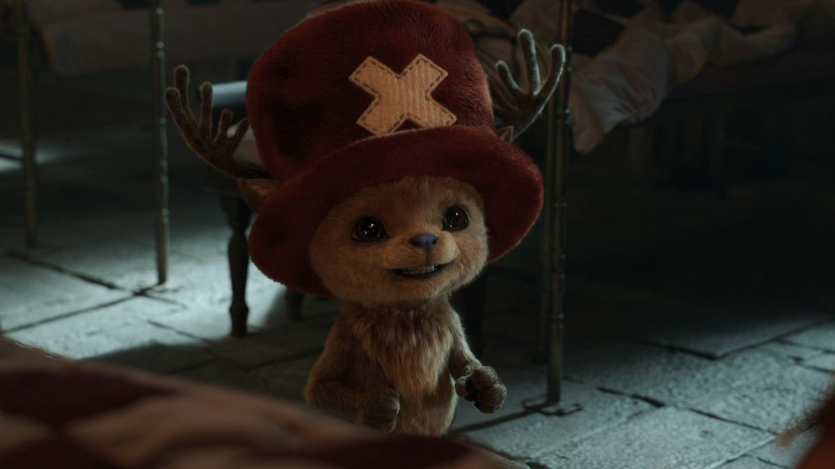 Tony Tony Chppper, a small, furry, cartoon-like character with antlers and a big red hat stands smiling indoors on a stone floor, with beds in the background, in a cozy dimly lit room.