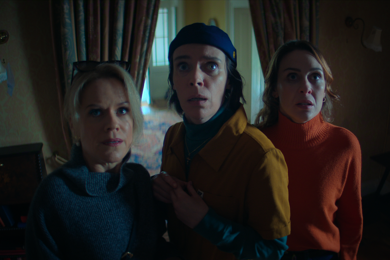 Three people indoors, standing close together and looking ahead with expressions of shock or concern, in a dimly lit room with curtains and vintage decor in the background.