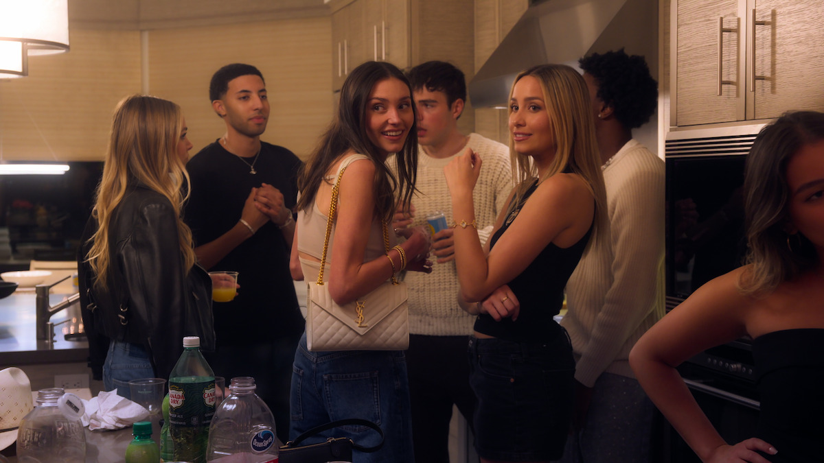 Alexie and Emilie at Jemma’s party. A brunette woman and blonde woman are standing amongst a group of friends in a kitchen.