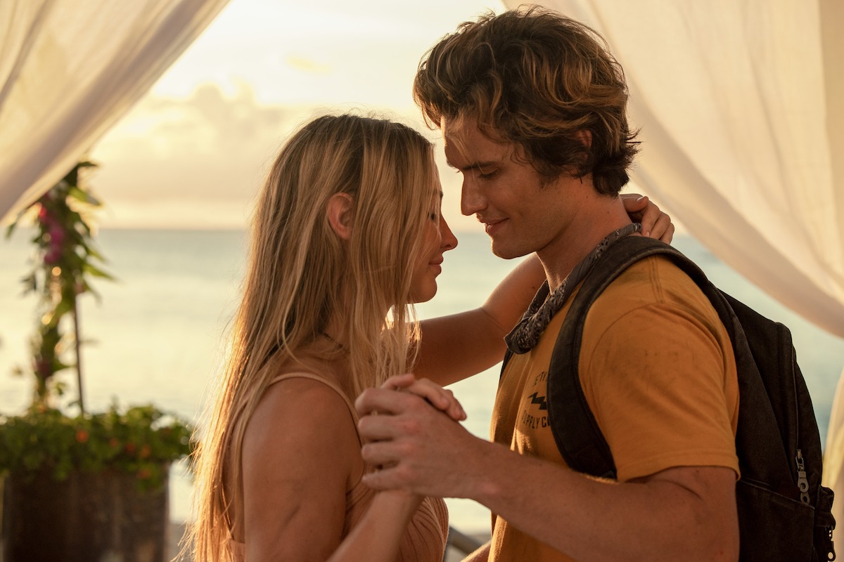Um casal jovem em pé e de mãos dadas se entreolha compartilhando um momento de intimidade, sob um tecido branco esvoaçante em uma praia ao pôr do sol. O cenário é romântico e aconchegante, com luz suave e o mar ao fundo.