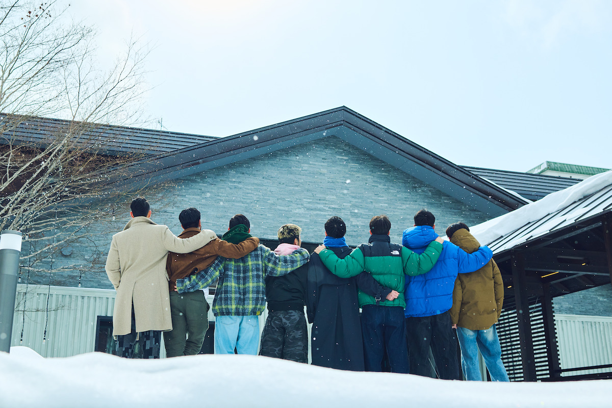 A group of people stand arm in arm outdoors in winter coats, facing away from the camera toward a snow-covered building under a bright sky. Snow is on the ground and bare trees are visible.