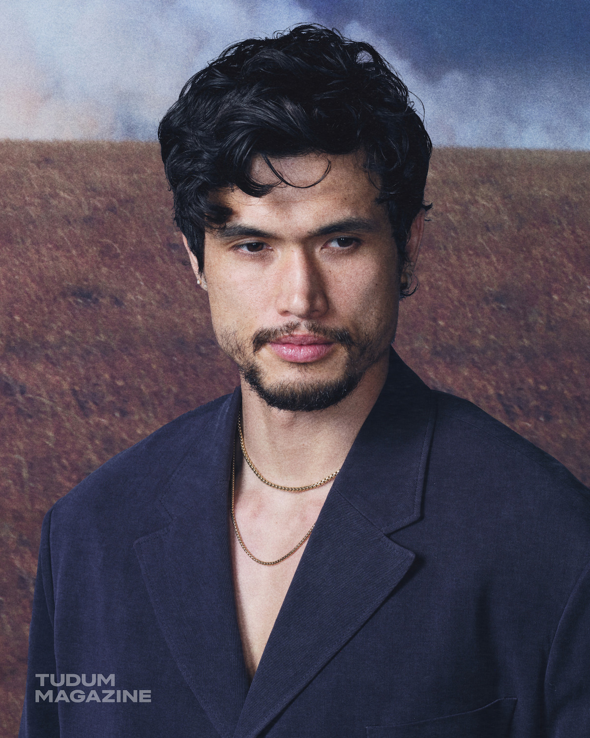 A man with dark hair and a beard wearing a navy blazer and gold chains stands in front of a blurred outdoor landscape with cloudy sky.