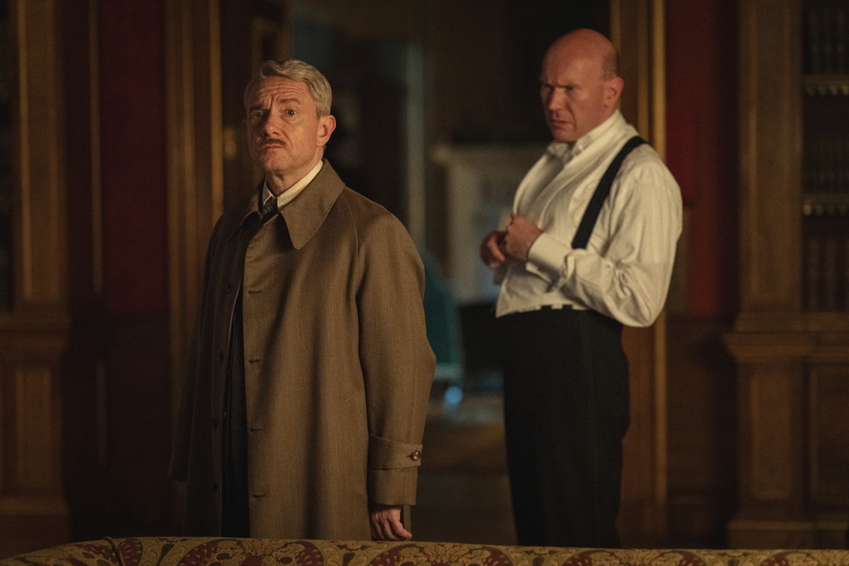 Two men stand in an elegant, dimly lit room with wood paneling and bookshelves; one wears a long coat and looks serious while the other adjusts his suspenders, adding a sense of tension to the scene.