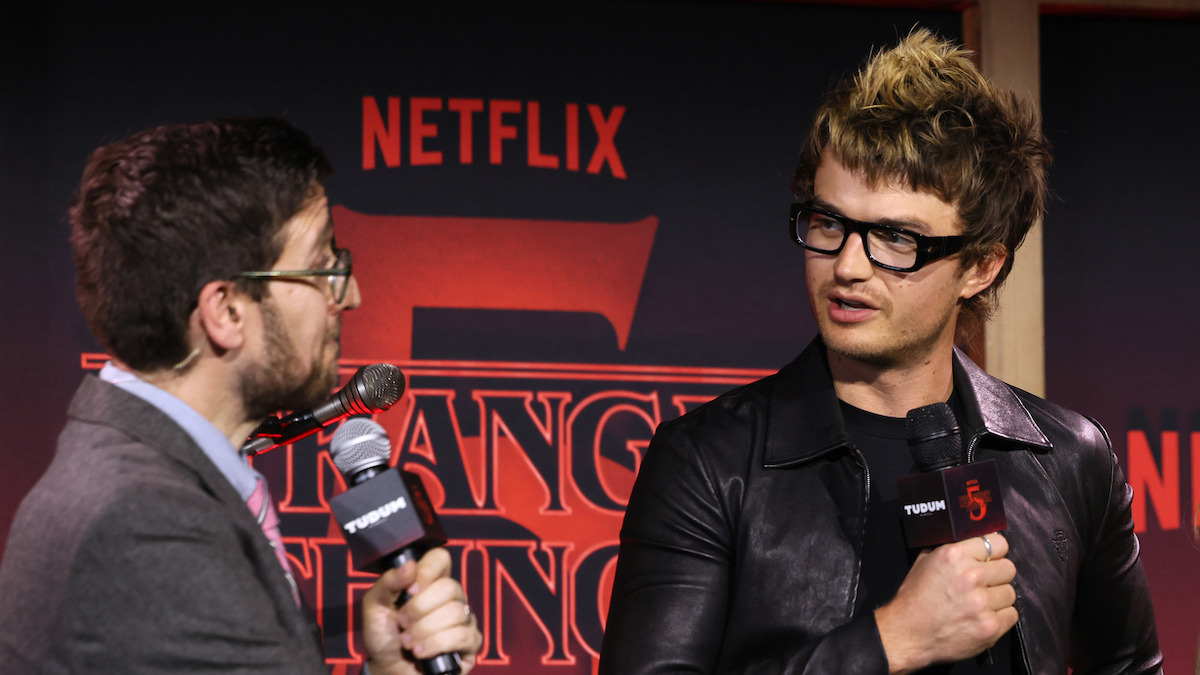 Josh Horowitz and Joe Keery (Steve Harrington) speaking into microphones at Netflix's Stranger Things 5 Premiere, with one in a suit and the other in a leather jacket, both appearing engaged and focused in a lively setting.