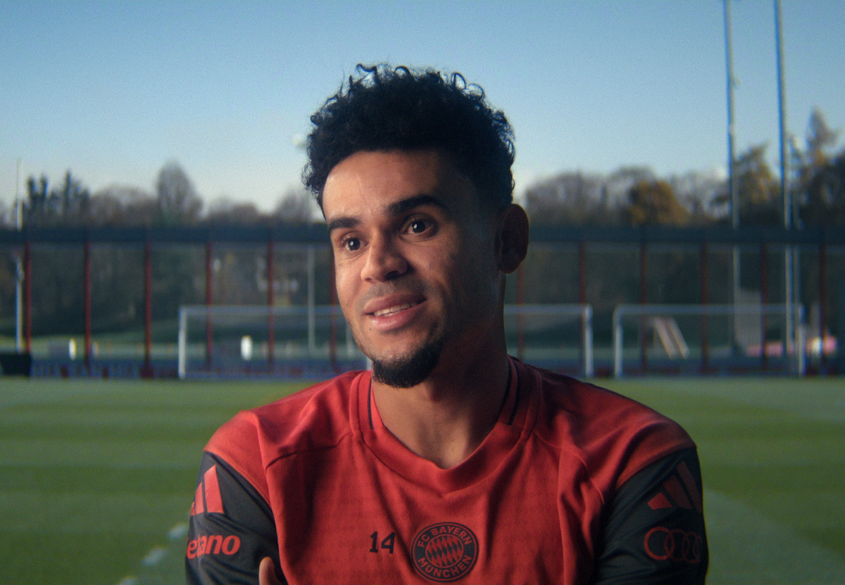A male soccer player with short curly hair and a trimmed beard, wearing a red training jersey, sits on a field with goalposts, outdoor training equipment, and trees in the background on a clear day.