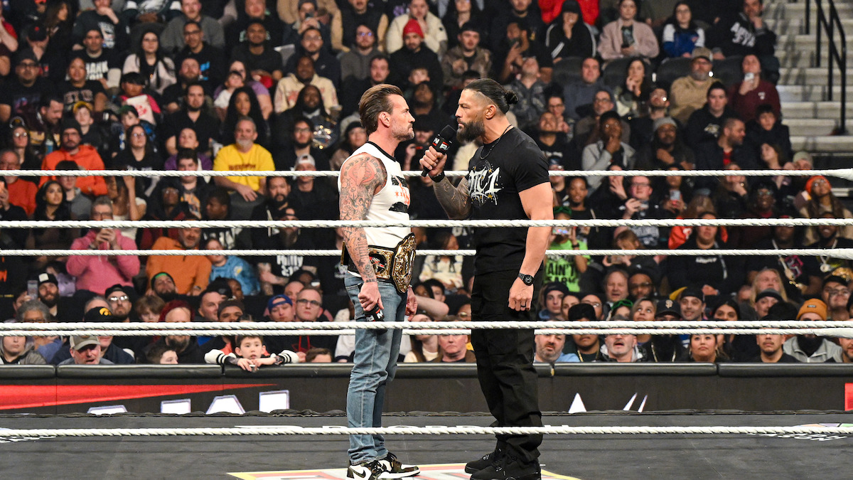 Two wrestlers face off in a packed arena wrestling ring, one holding a championship belt and the other speaking into a microphone as a large audience watches the dramatic confrontation.