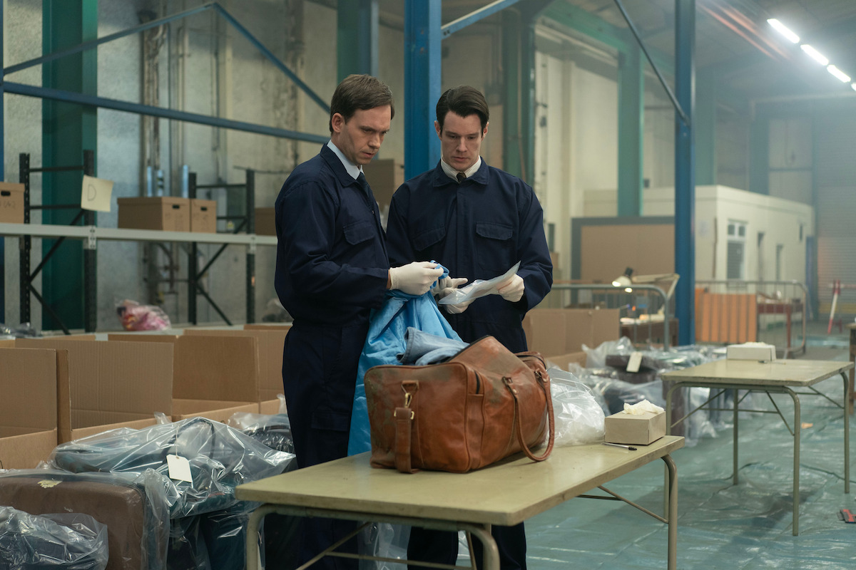 Two men in dark uniforms and gloves examine evidence from a bag in an industrial warehouse setting, surrounded by boxes, tables, and plastic-wrapped items, suggesting a crime investigation or forensic analysis environment.