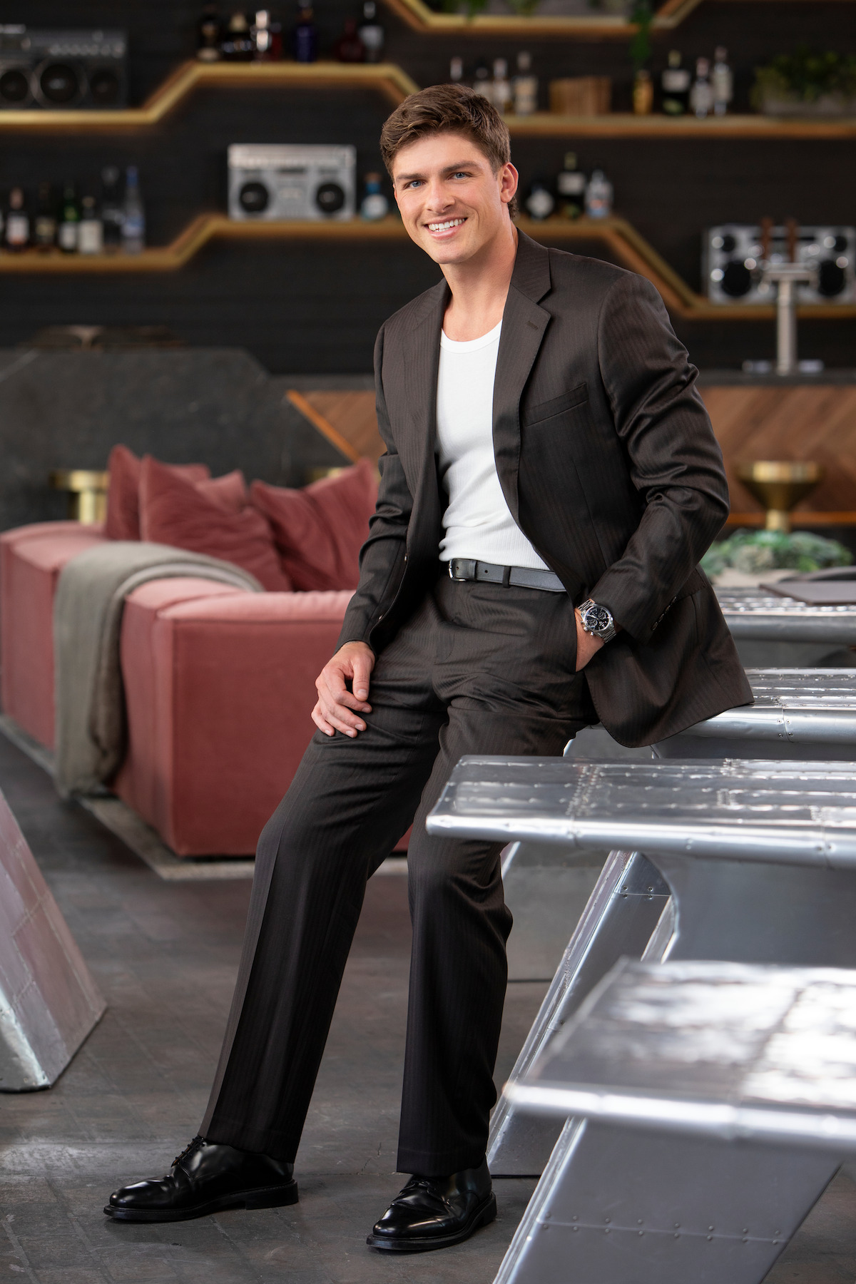 A smiling man in a dark suit and white shirt sits casually on a table in a modern lounge with stylish decor, metal surfaces, a cozy pink sofa, and shelves with bottles, creating a contemporary and relaxed atmosphere.