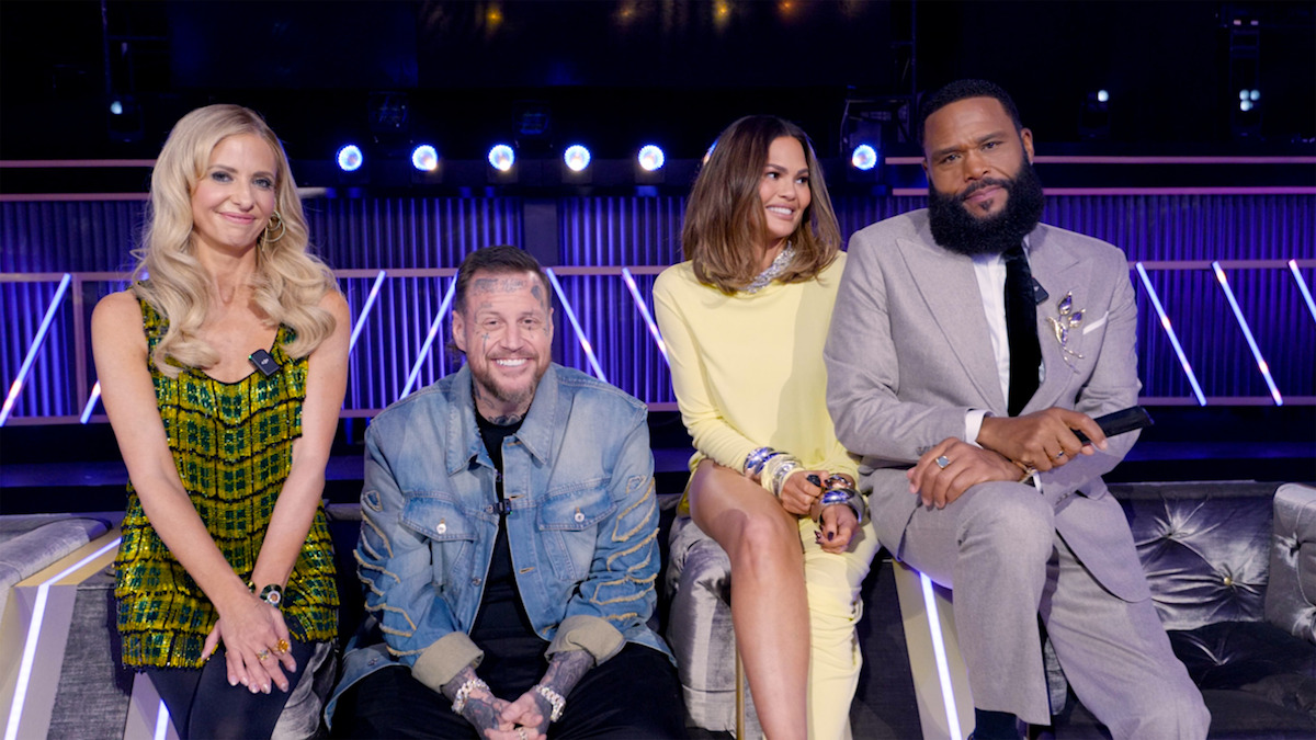 Four smiling people sit on a couch in a brightly lit studio, possibly a TV show set, with spotlights and a decorative background behind them.