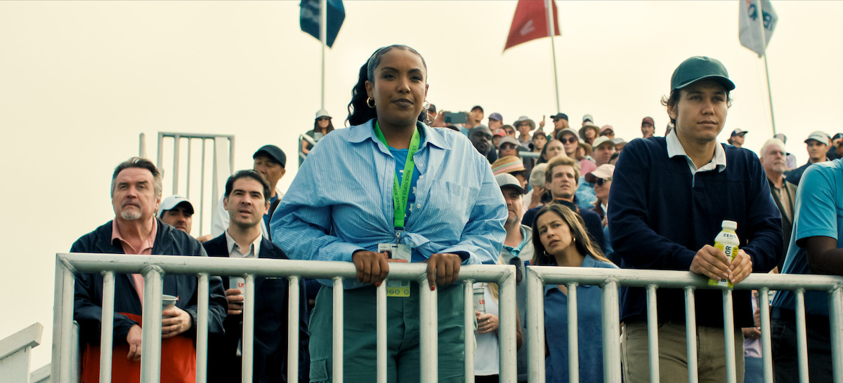 A crowd of people standing on stadium bleachers outdoors, some holding drinks, with flags in the background, watching an event attentively under a bright sky.