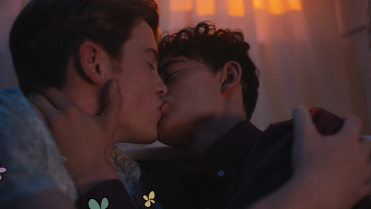 Two young men share an intimate kiss in a softly lit room with warm light filtering through curtains, creating a romantic and cozy atmosphere.