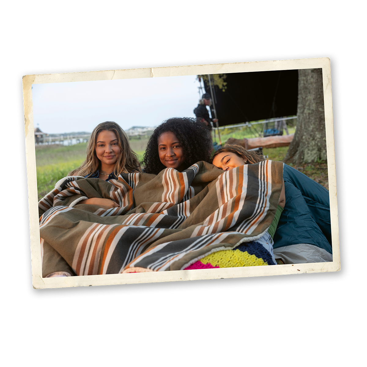 Three young women sitting outdoors wrapped in a striped blanket, relaxing together under a tree near grassy fields, with part of a crew visible in the background, creating a cozy and friendly atmosphere.