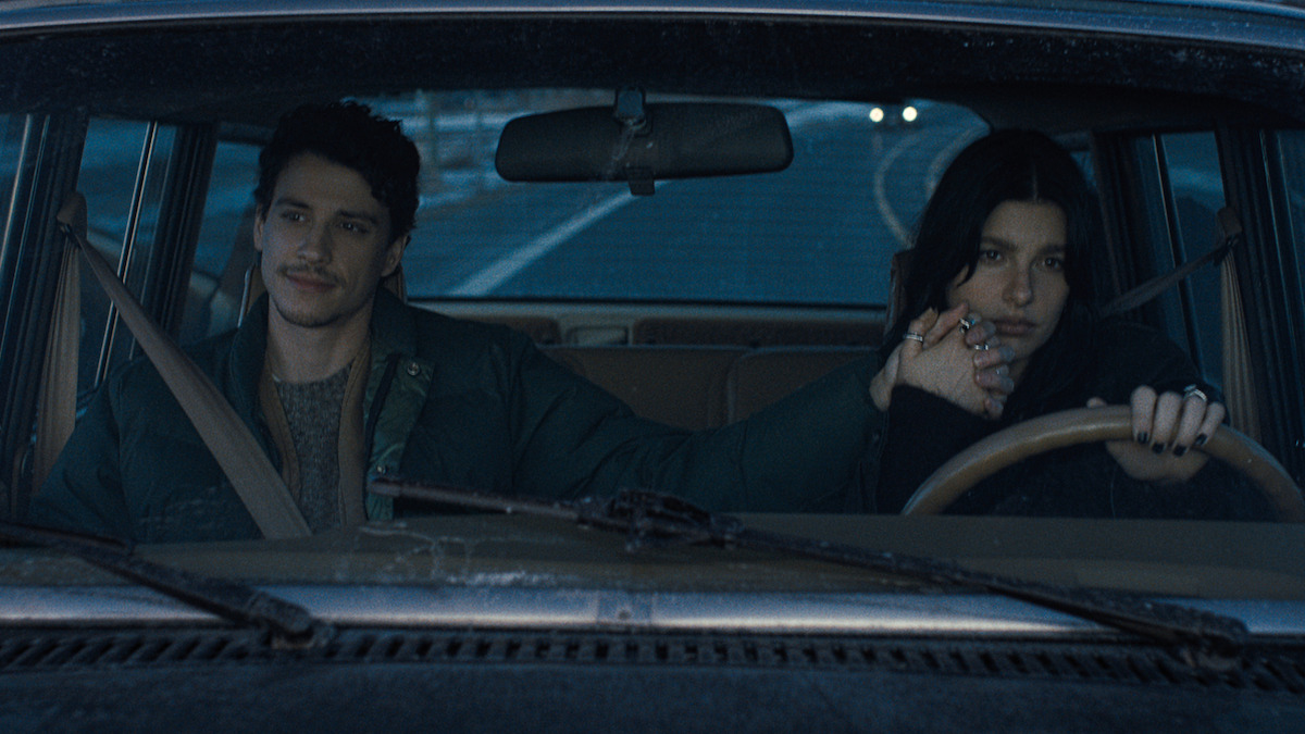 A man and woman sit in the front seats of a car at dusk, driving on a highway. The woman drives while holding hands with the man, both wearing seatbelts, expressing an intimate moment in a moody, quiet atmosphere.