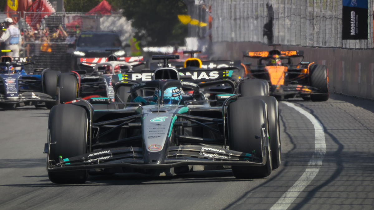 Formula 1 race cars speeding on a city street circuit, closely packed, with barriers and spectators in the background on a sunny day.