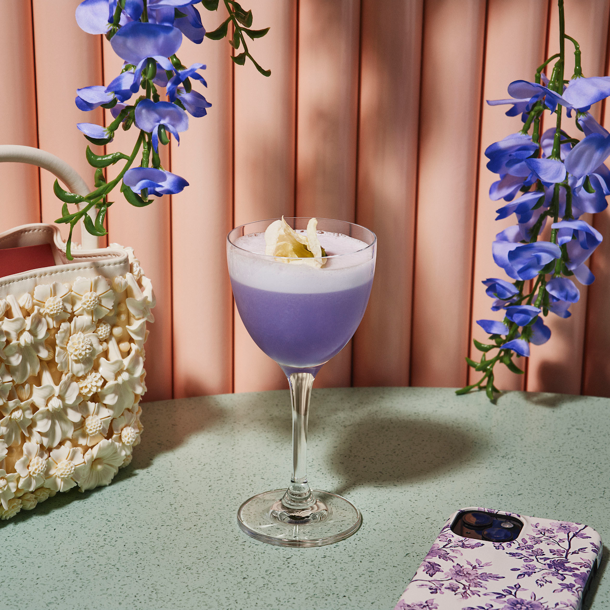 Lavender cocktail in a glass on a green table, surrounded by purple flowers, a decorative cream-colored handbag, and a floral phone case, set against a peach pleated backdrop.