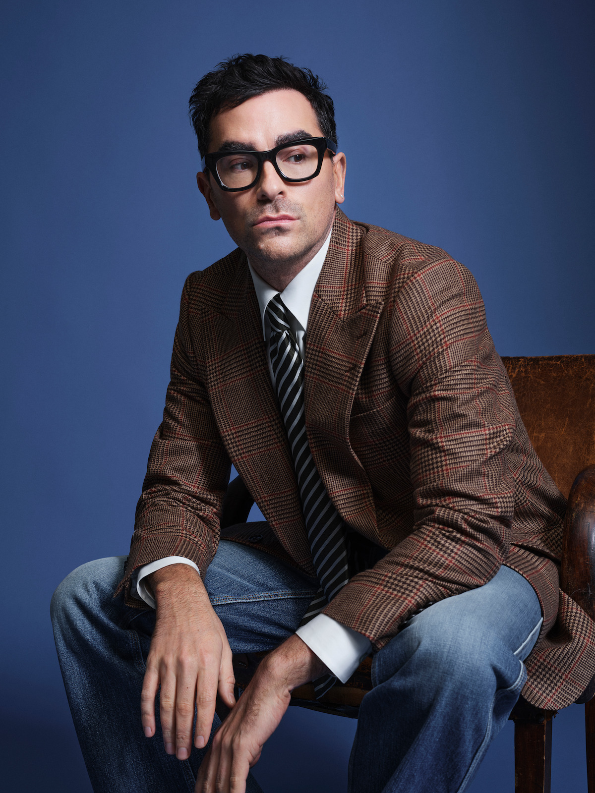 Dan Levy sits in a chair against a blue background.